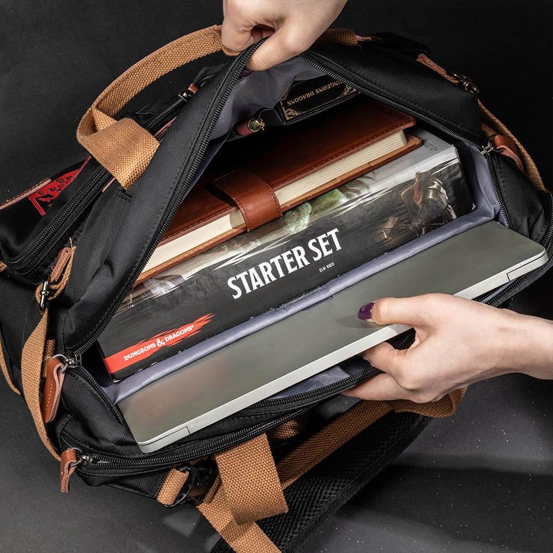 K&ouml;nnte beinhalten: Ein schwarzer Canvas-Rucksack mit braunen Lederelementen und einer Rei&szlig;verschluss&ouml;ffnung oben. Der Rucksack ist gef&uuml;llt mit B&uuml;chern, einem Laptop und einer Brettspielbox mit der Aufschrift "Starter Set" und "Dungeons & Dragons".