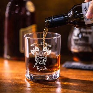 May include: A clear rocks glass with the phrase "THIS IS HOW I ROLL" and a graphic of two crossed axes and a D20 die. Amber liquid is being poured into the glass from a bottle. The glass sits on a wooden surface.