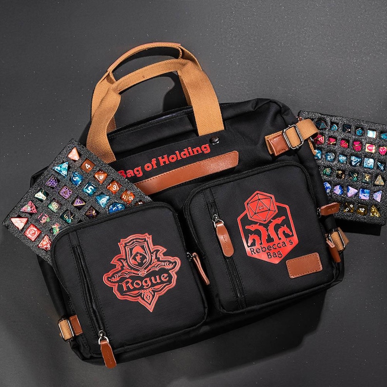 K&ouml;nnte beinhalten: Schwarze Canvas-Tasche mit braunen Lederelementen und roten Stickdetails. Die Tasche hat mehrere F&auml;cher und tr&auml;gt den Text "Bag of Holding" und "Rebecca's Bag".