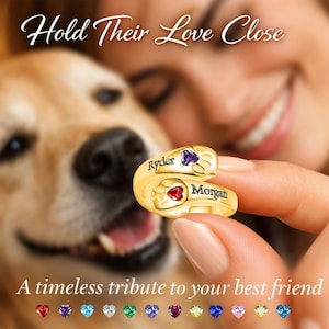 May include: A gold-toned wrap ring featuring two paw print motifs, each accented with a heart-shaped gemstone. The names Ryder and Morgan are engraved on the band. The text Hold Their Love Close and A timeless tribute to your best friend appears.