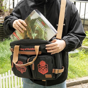 K&ouml;nnte beinhalten: Eine schwarze Canvas-Tasche mit braunen Lederakzenten und einem roten Logo, auf dem "Bag of Holding" steht. Die Tasche hat zwei Fronttaschen mit roten Logos, auf denen "Rogue" und "Rebecca's Bag" steht. Eine Person h&auml;lt eine Dungeons & Dragons Starter-Set-Box in der Hand, die sich teilweise in der Tasche befindet.