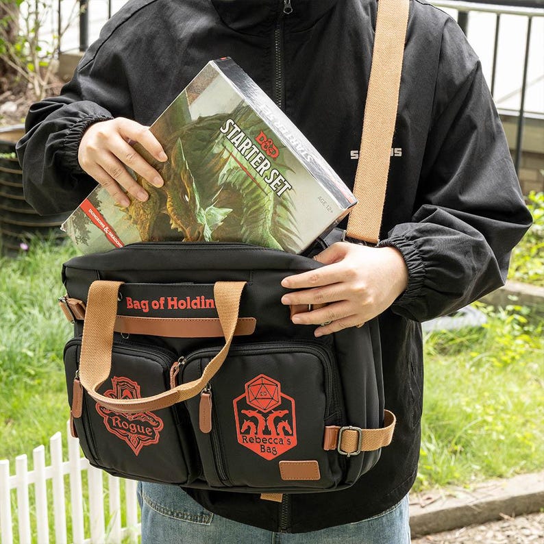 K&ouml;nnte beinhalten: Eine schwarze Canvas-Tasche mit braunen Lederakzenten und einem roten Logo, auf dem "Bag of Holding" steht. Die Tasche hat zwei Fronttaschen mit roten Logos, auf denen "Rogue" und "Rebecca's Bag" steht. Eine Person h&auml;lt eine Dungeons & Dragons Starter-Set-Box in der Hand, die sich teilweise in der Tasche befindet.