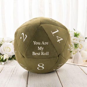 Puede incluir: Cojín en forma de dado d20 de color verde oliva con números blancos cosidos y la frase "You Are My Best Roll". El cojín está sobre una superficie de madera blanca con flores blancas en el fondo.