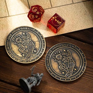 Puede incluir: Dos posavasos de metal de color dorado con un diseño en blanco y negro de una mano esquelética sosteniendo un dado. Los posavasos tienen el texto "What one hand gives" alrededor del borde.