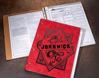May include: A red binder with the word "JOHNWICK" in black lettering, featuring a dragon and geometric design. The binder is open, revealing pages with printed text and a clear sheet protector. A brown folder is also open, displaying printed text.