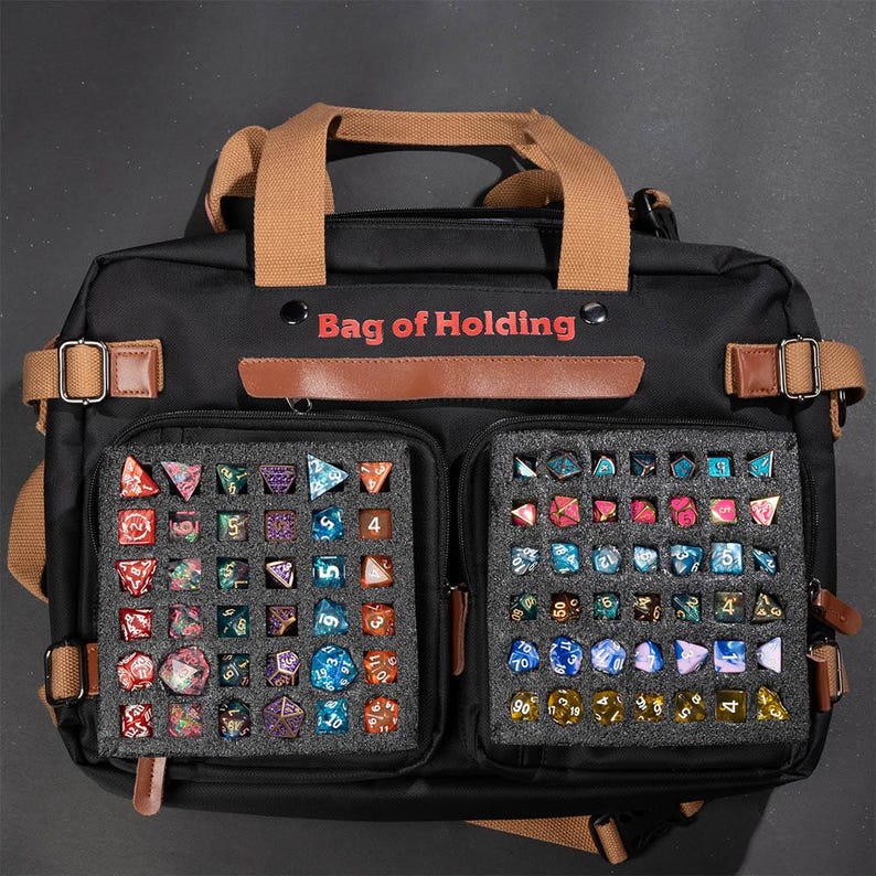 K&ouml;nnte beinhalten: Eine schwarze Canvas-Tasche mit braunen Lederakzenten und dem Aufdruck "Bag of Holding" auf der Vorderseite. Die Tasche hat zwei gro&szlig;e F&auml;cher mit Schaumstoffeinlagen zum Verstauen von W&uuml;rfeln. Die F&auml;cher sind mit einer Vielzahl von bunten W&uuml;rfeln gef&uuml;llt.