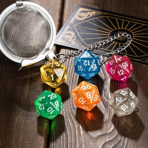 May include: A set of six colorful, multi-sided dice with white numbers on each side. The dice are attached to a silver tea strainer with a chain. The dice are on a wooden surface.