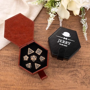 Puede incluir: Una caja hexagonal de cuero marrón para dados que contiene un juego de siete dados poliédricos de metal dorado con numeración negra. Al lado hay una caja hexagonal negra cerrada con un gráfico blanco de bombín, pajarita y el texto JERRY 04.06.2025.
