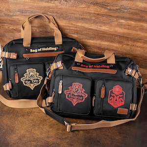 K&ouml;nnte beinhalten: Zwei schwarze Canvas-Taschen mit braunen Lederakzenten und Riemen. Die Taschen haben den Text "Bag of Holding" auf der Vorderseite. Eine Tasche hat ein goldenes Emblem mit dem Text "the DND" und die andere ein rotes Emblem mit dem Text "Rogue" und "Rebecca's Bag".