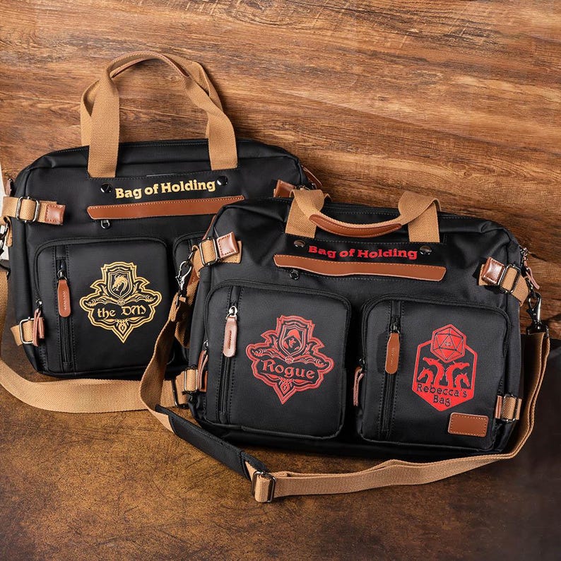 K&ouml;nnte beinhalten: Zwei schwarze Canvas-Taschen mit braunen Lederakzenten und Riemen. Die Taschen haben den Text "Bag of Holding" auf der Vorderseite. Eine Tasche hat ein goldenes Emblem mit dem Text "the DND" und die andere ein rotes Emblem mit dem Text "Rogue" und "Rebecca's Bag".
