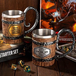 Könnte beinhalten: Zwei braune und metallene Becher mit Holzmaserung. Die Becher haben ein Edelstahl-Innenleben und einen Griff. Ein Becher trägt den Text "World Okayest Dungeon Master". Der andere Becher trägt den Text "Because I'm the DM That's why."