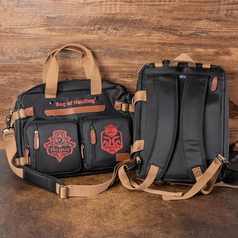 K&ouml;nnte beinhalten: Zwei schwarze Canvas-Taschen mit braunen Lederelementen. Die kleinere Tasche hat den Text "Bag of Holding" und "Rebecca's Bag" aufgedruckt. Die gr&ouml;&szlig;ere Tasche hat den Text "Rogue" aufgedruckt. Beide Taschen haben einen braunen Riemen.