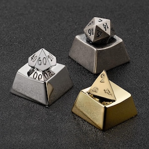 May include: Three metal dice keycaps in silver, gunmetal, and gold. Each keycap has a different dice shape and number arrangement. The keycaps are designed to fit on a mechanical keyboard and are displayed on a dark surface.