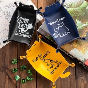 May include: Three leather dice trays in black, navy blue, and yellow. Each tray features a cat design and text: "Clickity Clackity I roll to Attackitty" or "Mother Fluffer". Green dice are scattered around the yellow tray.