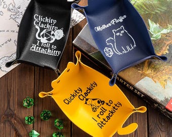 May include: Three leather dice trays in black, navy blue, and yellow. Each tray features a cat design and text: "Clickity Clackity I roll to Attackitty" or "Mother Fluffer". Green dice are scattered around the yellow tray.