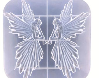 May include: A clear silicone mold featuring two detailed fairy designs. Each fairy has outstretched wings and is holding an object. The mold is square with a grid pattern, designed for crafting projects. The fairies are in a symmetrical arrangement.