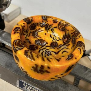 May include: A close-up of a small, orange and black resin bowl with a swirling pattern. The bowl is sitting on a metal surface with a "Lumber Jack" logo.