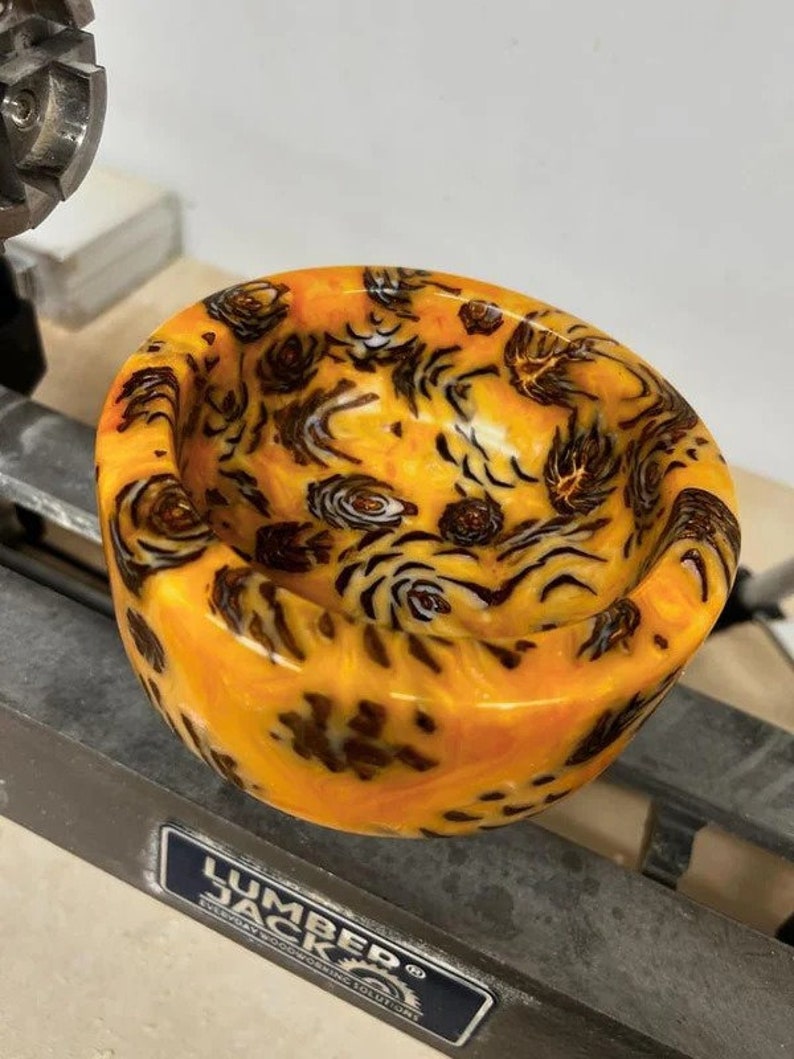 May include: A close-up of a small, orange and black resin bowl with a swirling pattern. The bowl is sitting on a metal surface with a "Lumber Jack" logo.
