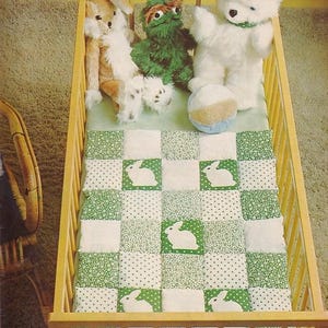May include: A wooden baby crib with a green and white patchwork quilt featuring bunny appliques. The crib contains stuffed animals, including a white teddy bear, a brown bunny, and a green monster.