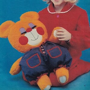 Può includere: Un orsacchiotto arancione di peluche con un viso sorridente, guance rosse e occhi viola chiusi. L'orso indossa una tuta blu a pois con finiture rosse. L'orso è tenuto da una persona che indossa un completo rosso.