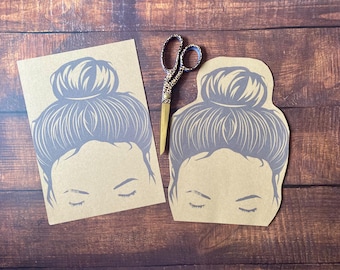 May include: Two brown paper cutouts of a woman with a bun hairstyle. The cutouts are on a wooden surface.
