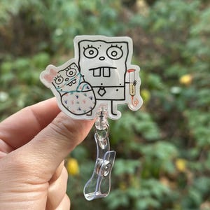 May include: A clear, cartoon-style badge reel featuring a black and white drawing of a character holding another character. The badge reel has a clear plastic clip and retractable cord.