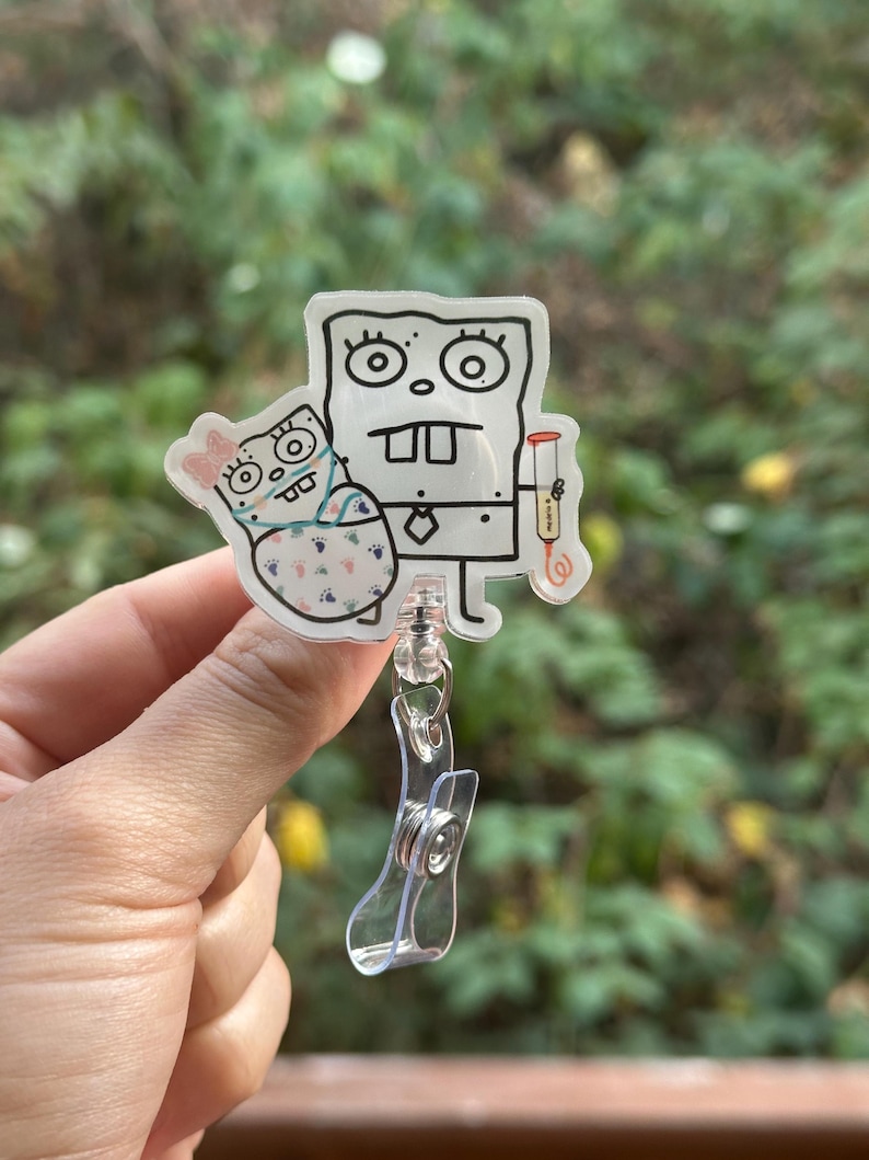 May include: A clear, cartoon-style badge reel featuring a black and white drawing of a character holding another character. The badge reel has a clear plastic clip and retractable cord.