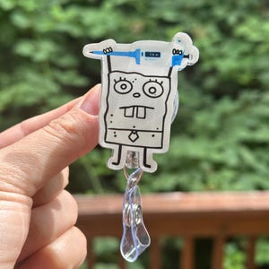 May include: A clear acrylic badge reel featuring a cartoon character with a syringe design. The character is white with black outlines and has a square shape. The badge reel has a clear plastic clip and a retractable cord.