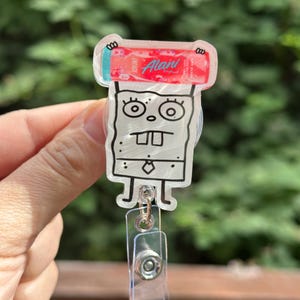 May include: A clear acrylic badge reel featuring a cartoon character with a red and pink energy drink. The badge reel has a clear plastic clip and a retractable cord.