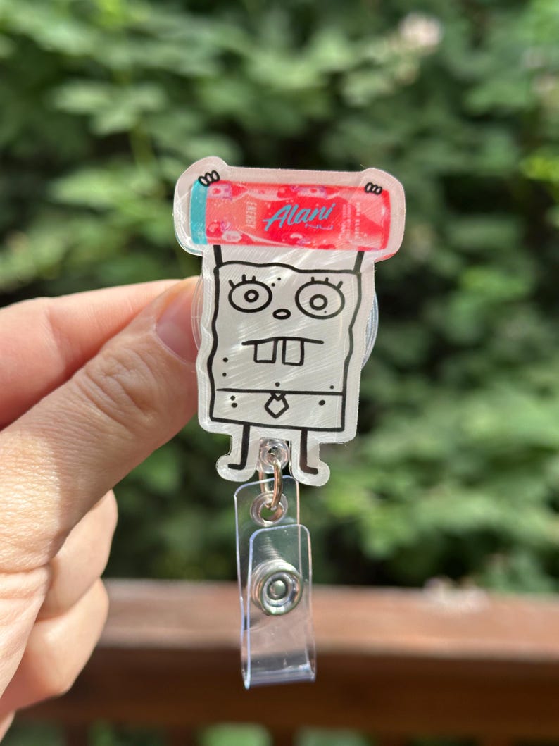 May include: A clear acrylic badge reel featuring a cartoon character with a red and pink energy drink. The badge reel has a clear plastic clip and a retractable cord.