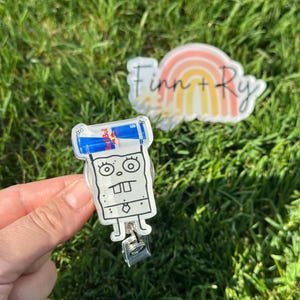 Peut inclure: Un porte-badge en acrylique transparent avec un personnage de dessin anim&eacute; tenant deux canettes de boisson &eacute;nergisante Red Bull. Le porte-badge a un clip argent&eacute; et un dos en plastique transparent. Un autocollant avec un arc-en-ciel et le texte "Finn + Ry" est &eacute;galement visible.