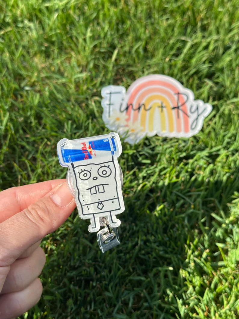 Peut inclure: Un porte-badge en acrylique transparent avec un personnage de dessin anim&eacute; tenant deux canettes de boisson &eacute;nergisante Red Bull. Le porte-badge a un clip argent&eacute; et un dos en plastique transparent. Un autocollant avec un arc-en-ciel et le texte "Finn + Ry" est &eacute;galement visible.