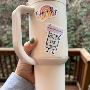 May include: A cream-colored insulated tumbler with a handle, adorned with two stickers. One sticker features the text "Finn + Ry" above a rainbow design. The other sticker is a cartoon character. The tumbler is held against a blurred outdoor background.