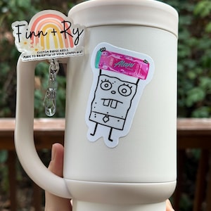 May include: A white tumbler with a handle, adorned with two stickers. One sticker features a rainbow design with the text "Finn + Ry" and "CUSTOM BADGE REELS MADE TO BRIGHTEN UP YOUR WORKPLACE!". The other sticker is a cartoon character.