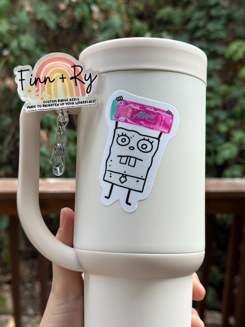 May include: A white tumbler with a handle, adorned with two stickers. One sticker features a rainbow design with the text "Finn + Ry" and "CUSTOM BADGE REELS MADE TO BRIGHTEN UP YOUR WORKPLACE!". The other sticker is a cartoon character.