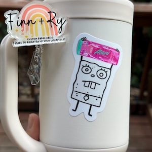 May include: A white mug with two stickers and a custom badge reel. The badge reel has a rainbow design and the text "Finn + Ry". The other sticker is a cartoon character holding a pink energy drink.