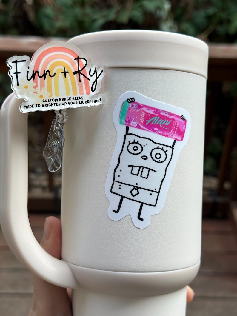 May include: A white mug with two stickers and a custom badge reel. The badge reel has a rainbow design and the text "Finn + Ry". The other sticker is a cartoon character holding a pink energy drink.