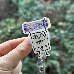 May include: A clear acrylic badge reel featuring a cartoon character with a square body, large eyes, and buck teeth. The reel has a purple and blue design with the word "Alani" on top. The reel is held by a clear plastic clip.