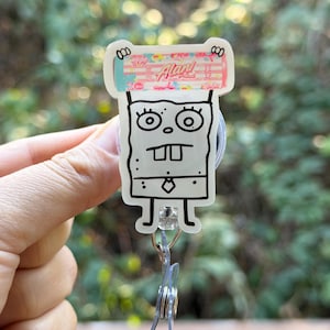 May include: A clear badge reel featuring a cartoon character design. The character is a yellow square with a black outline, large eyes, and a tie. The top of the reel has the word "Alani" in pink and red. A clear plastic clip and a metal ring are attached.