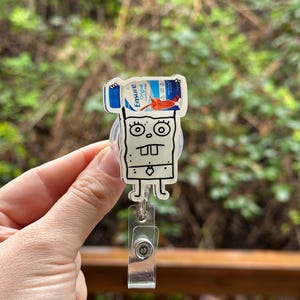 May include: A white cartoon character with a black outline holding a blue and white Ensure Original Nutrition Shake bottle above its head. The character is attached to a clear plastic retractable badge holder.