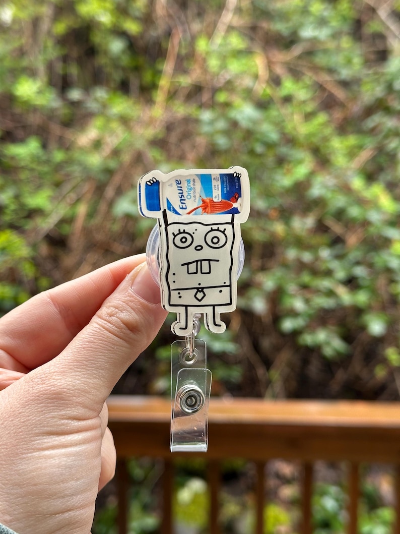 May include: A white cartoon character with a black outline holding a blue and white Ensure Original Nutrition Shake bottle above its head. The character is attached to a clear plastic retractable badge holder.