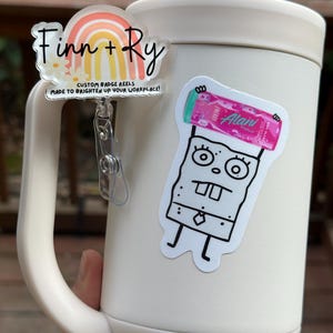 May include: A white tumbler with a handle, featuring two stickers and a badge reel. The badge reel has a rainbow design with the names "Finn + Ry" and the text "CUSTOM BADGE REELS MADE TO BRIGHTEN UP YOUR WORKPLACE!". One sticker is a cartoon character, and the other is a pink energy drink.