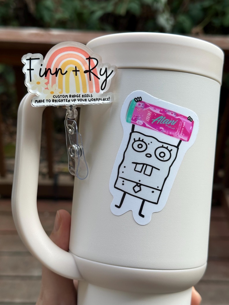 May include: A white tumbler with a handle, featuring two stickers and a badge reel. The badge reel has a rainbow design with the names "Finn + Ry" and the text "CUSTOM BADGE REELS MADE TO BRIGHTEN UP YOUR WORKPLACE!". One sticker is a cartoon character, and the other is a pink energy drink.