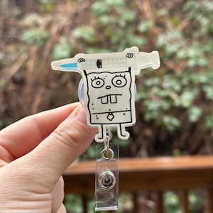 May include: A clear plastic badge holder with a cartoon character holding a syringe. The character is white with black outlines and has a tie. The badge holder has a clear retractable reel.