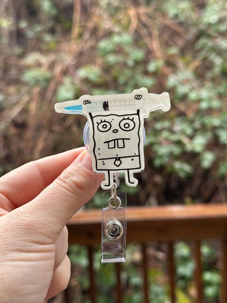 May include: A clear plastic badge holder with a cartoon character holding a syringe. The character is white with black outlines and has a tie. The badge holder has a clear retractable reel.