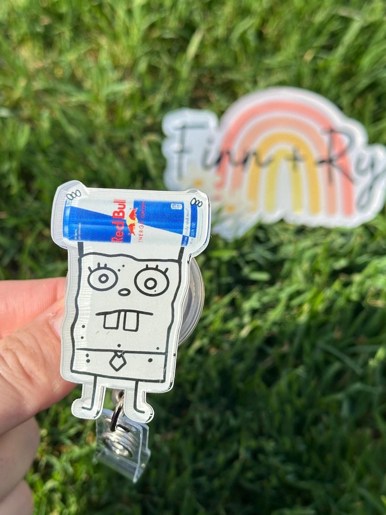 Peut inclure: Un porte-badge en acrylique transparent avec un dessin de dessin anim&eacute; de Bob l'&eacute;ponge tenant une canette de boisson &eacute;nergisante Red Bull. Le porte-badge a un cordon r&eacute;tractable et un clip. Un motif arc-en-ciel avec le texte "Finn + Ry" est en arri&egrave;re-plan.
