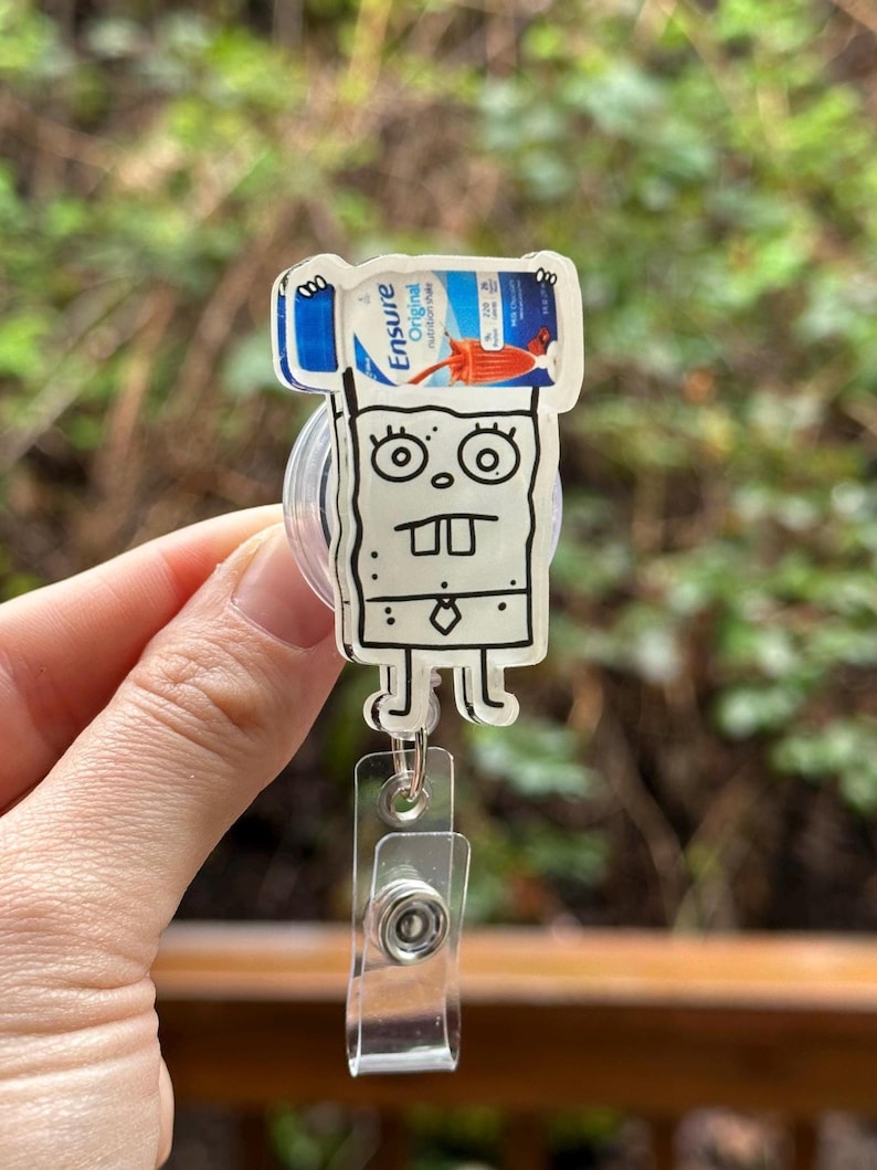 May include: A cartoon character resembling SpongeBob SquarePants holding a bottle of Ensure Original Nutrition Shake. The character is attached to a clear plastic retractable badge holder.