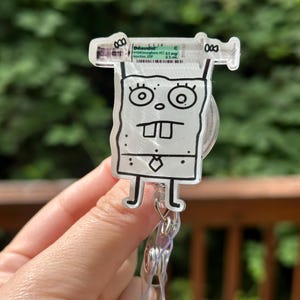 Op de afbeelding: Een doorzichtige, cartoonachtige badge reel met een spuitontwerp en de tekst "Dilaudid" en een SpongeBob SquarePants-personage. De badge reel heeft een doorzichtige plastic clip en een intrekbaar koord.