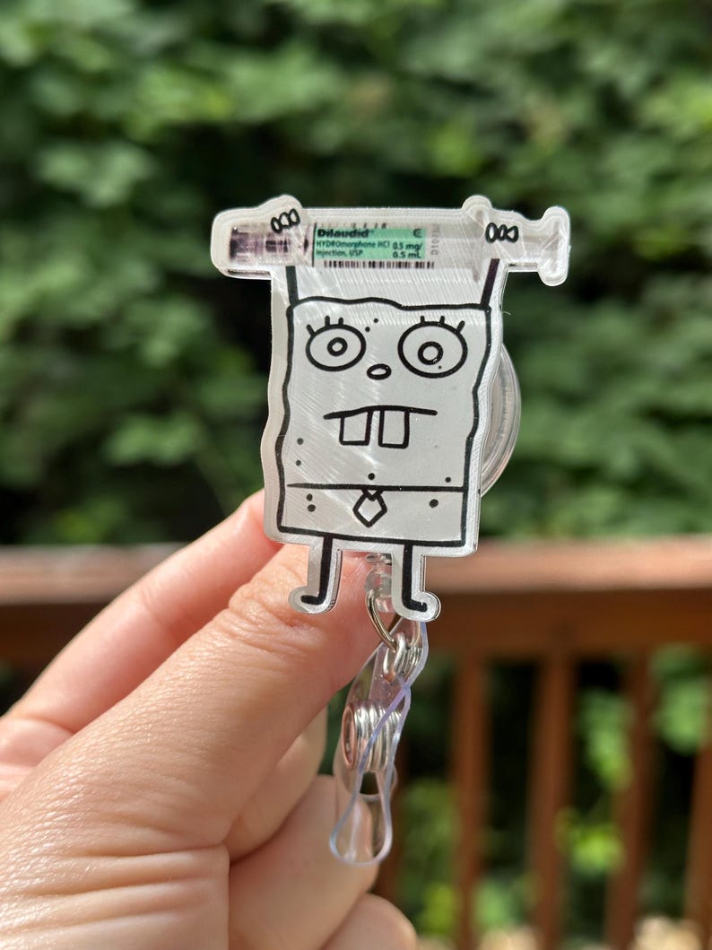 Op de afbeelding: Een doorzichtige, cartoonachtige badge reel met een spuitontwerp en de tekst "Dilaudid" en een SpongeBob SquarePants-personage. De badge reel heeft een doorzichtige plastic clip en een intrekbaar koord.