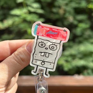 May include: A clear acrylic badge reel featuring a cartoon character design. The character has a square body, large eyes, and a tie. The badge reel has a red and white design with the text "Alani".